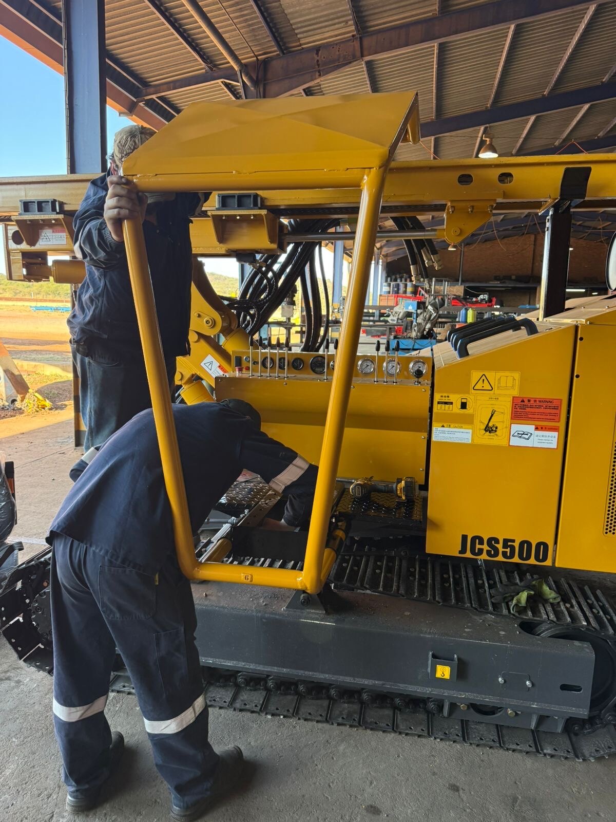 Two technicians fitting the yellow sun canopy over the operator station of a JCS500, with JCS500 branding and control gauges visible