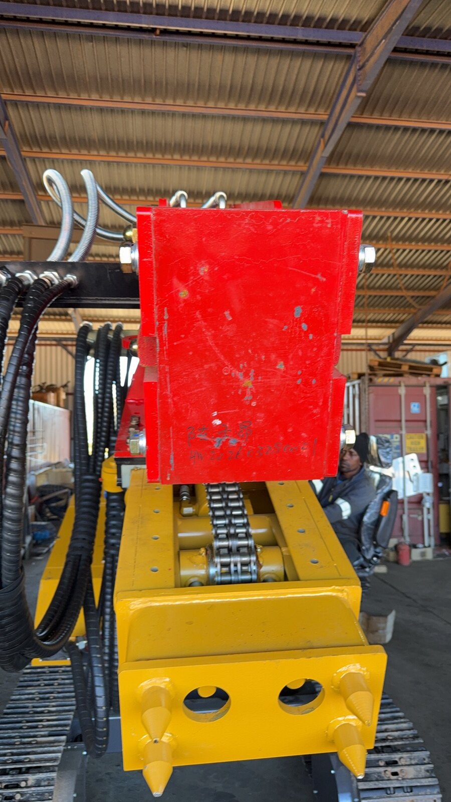 Bottom of the feed mast showing the red power-head carriage on the feed chain and a yellow bolt-on foot with bush-mount holes and ground spikes