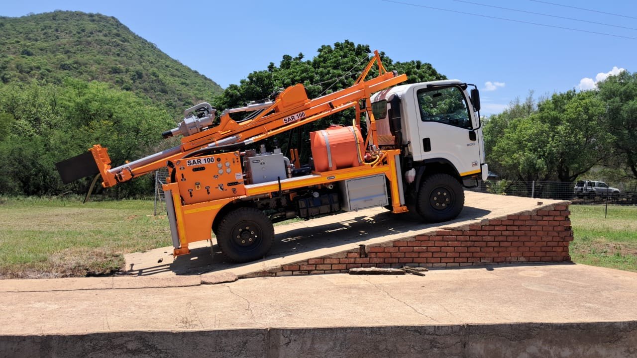 Side profile of an orange SAR100 drilling rig on a compact truck, mast stowed, parked on a raised brick ramp in the bushveld