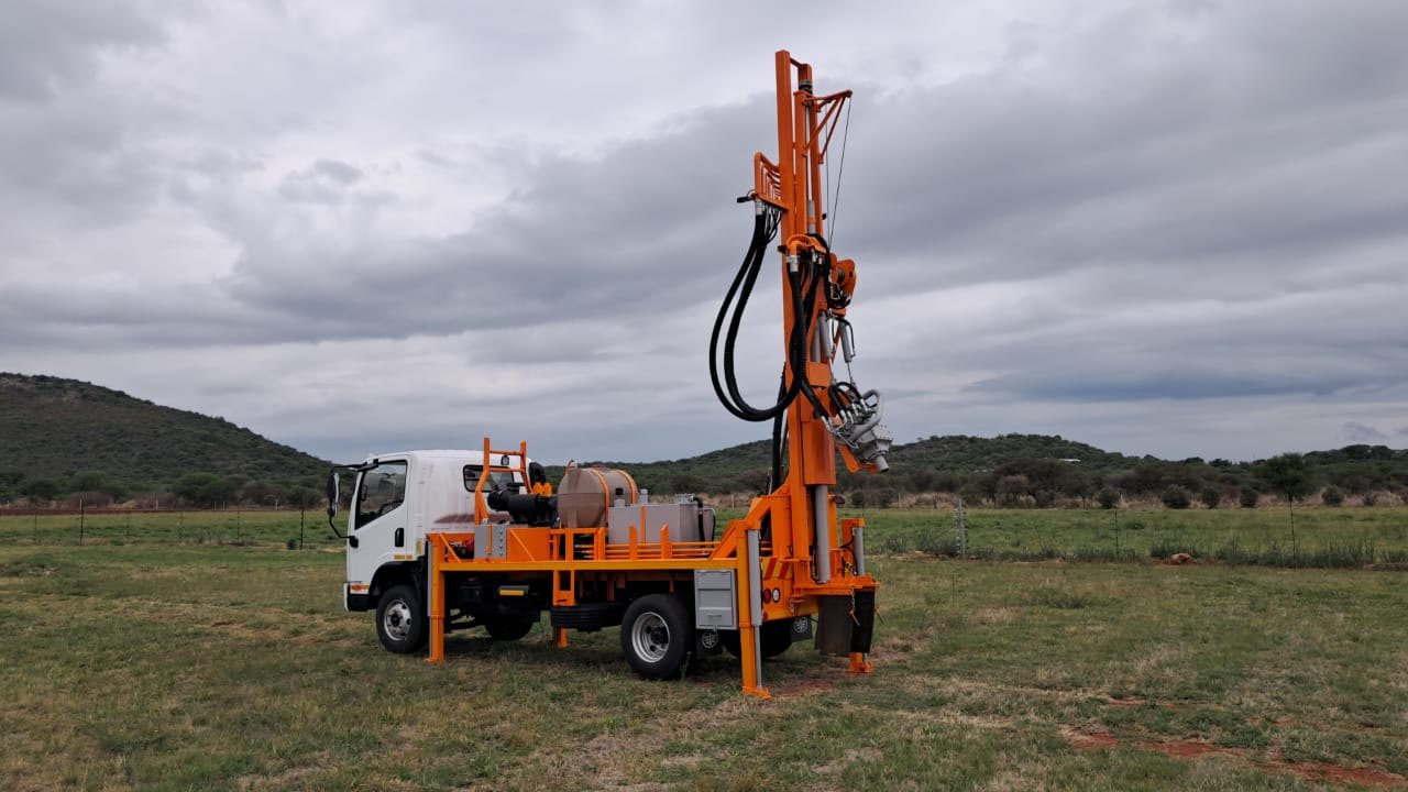 Orange SAR100 drilling rig set up in open veld with its mast raised vertical, outriggers down, under a stormy sky
