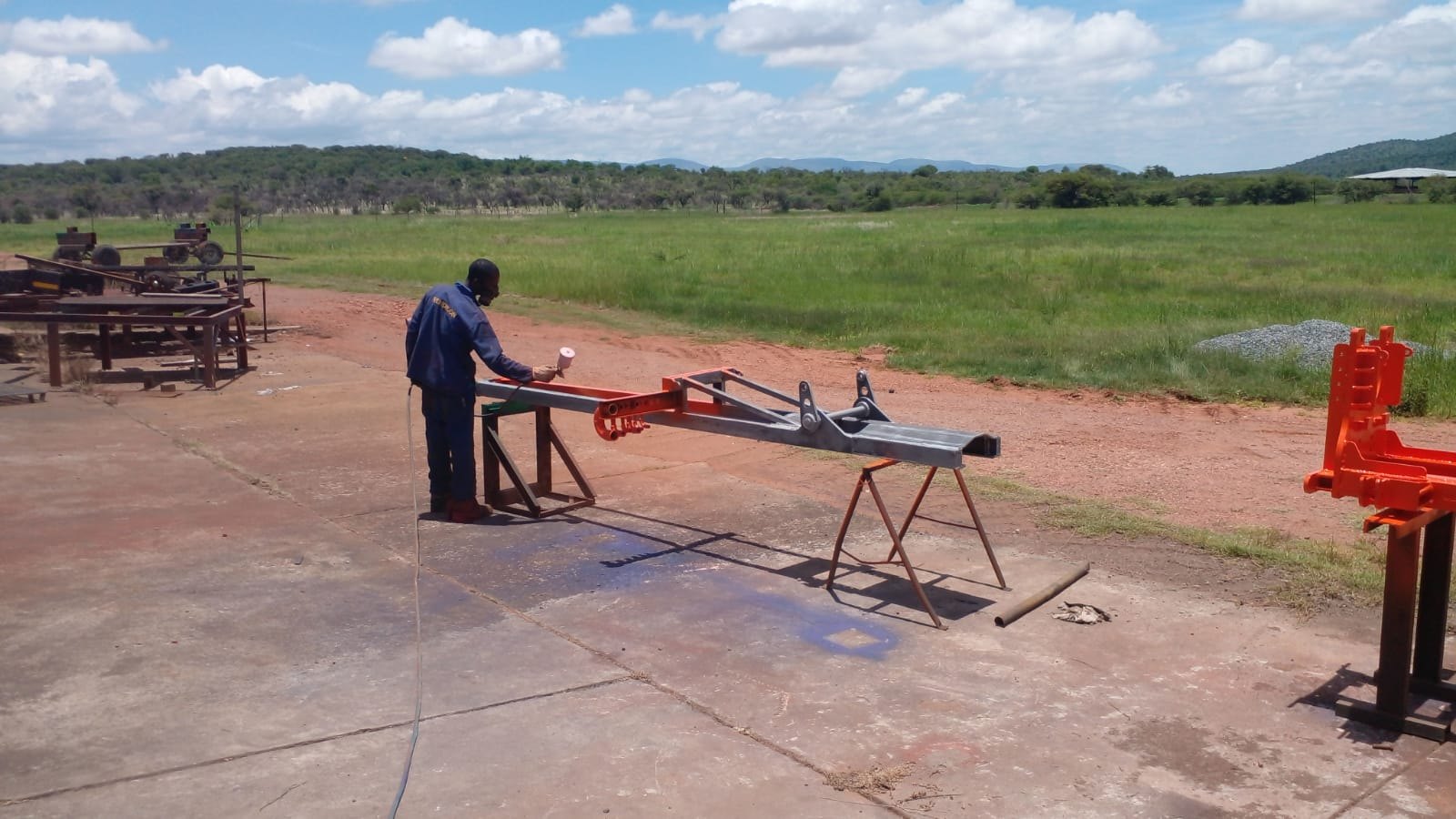 Painter spraying a SAR100 mast and feed beam on trestles outside the workshop, bare steel turning orange